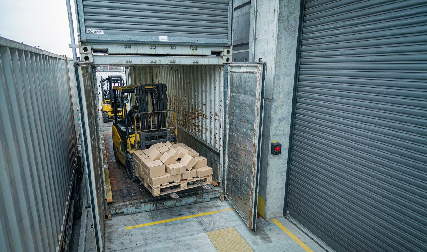 Forklift loading palletised export cartons into a shipping container at a loading dock